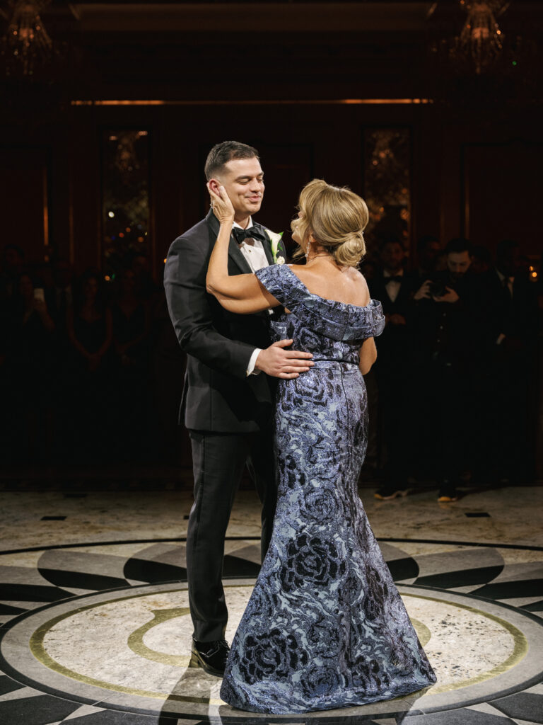 Emotional mother of the groom in a luxury off-the-shoulder metallic blue sequin gown dancing with her son in a classic black tuxedo