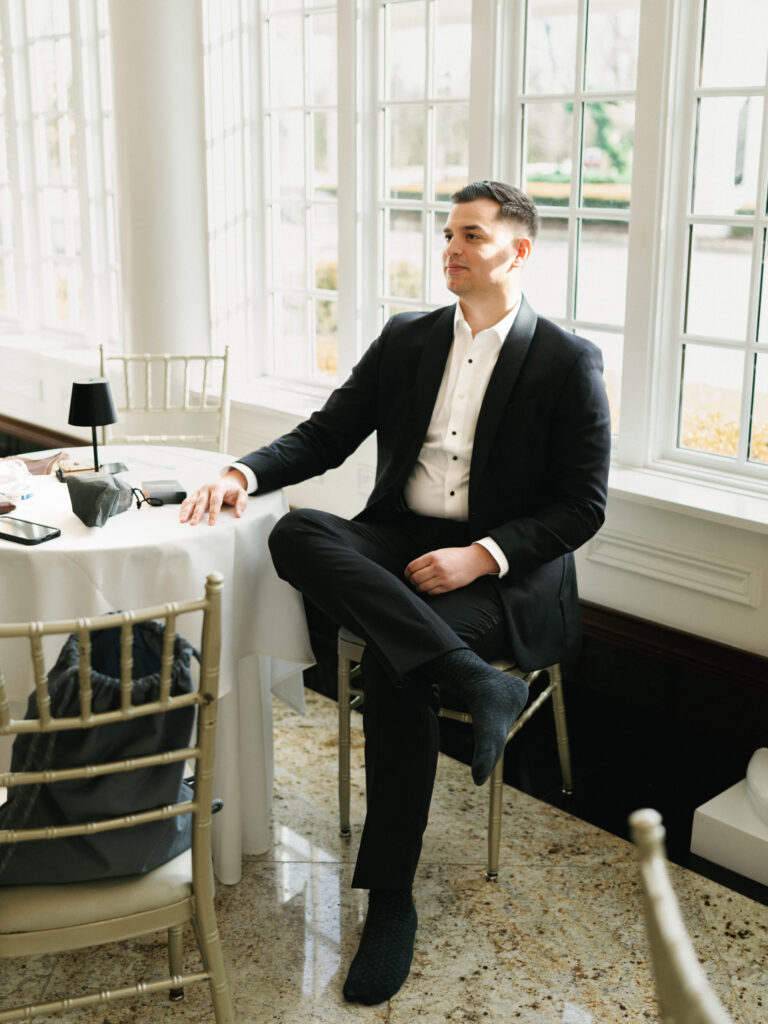Elegant groom portrait featuring a classic black tuxedo with satin lapels in a sunlit luxury wedding venue reception hall