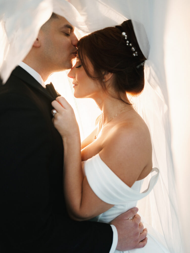 Romantic bridal portrait of a couple sharing an intimate moment under a cathedral veil, showcasing an elegant off-the-shoulder wedding gown