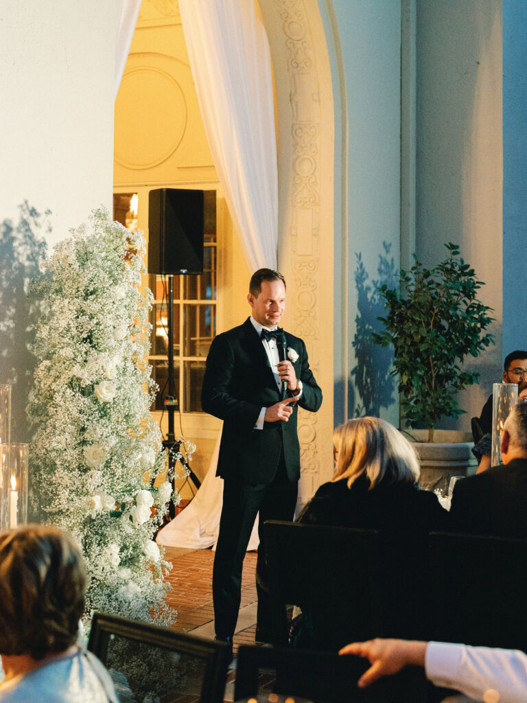 A man in a black tuxedo giving a speech during an intimate indoor wedding reception with warm evening lighting