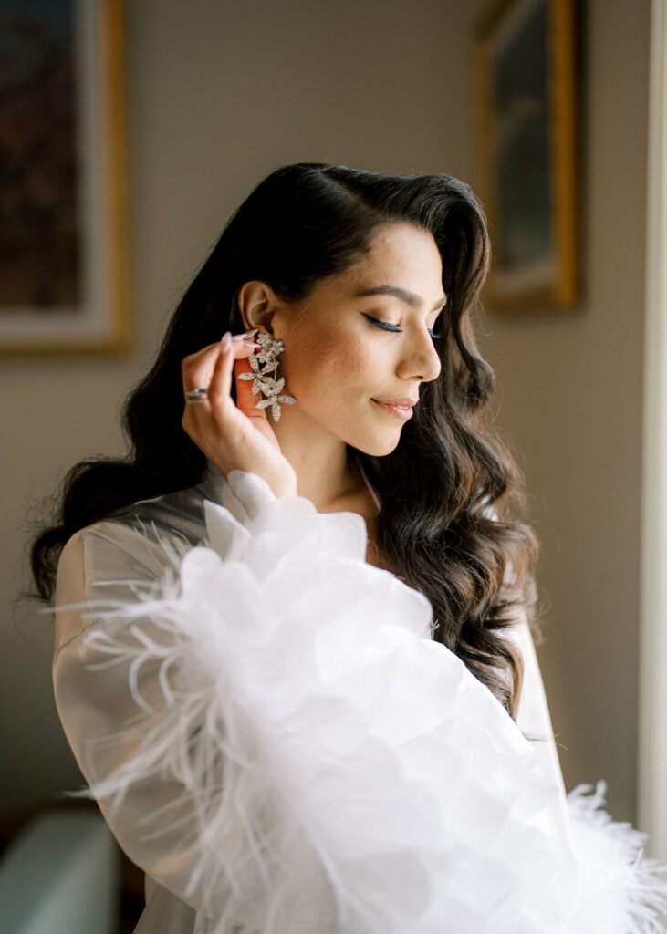 Elegant bride getting ready for a luxury destination wedding in France, putting on statement diamond earrings in a couture silk robe with feathers