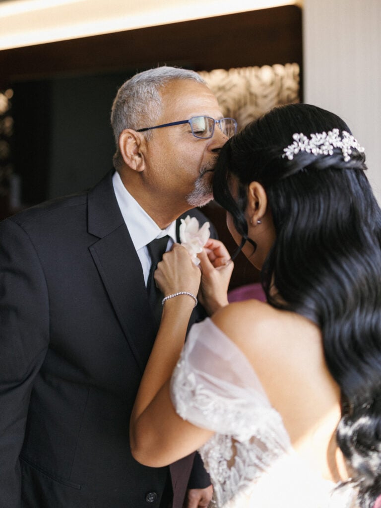 Emotional father of the bride kissing his daughter's forehead while getting ready and adjusting his boutonniere before the wedding ceremony