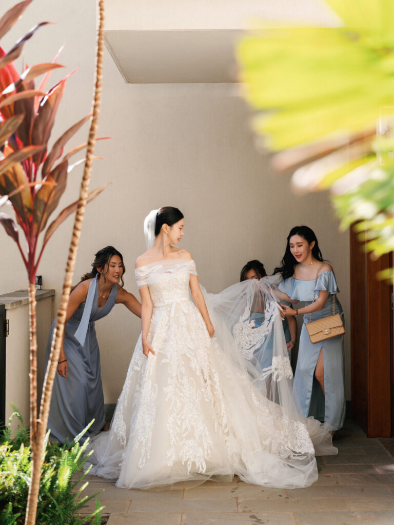 Bride in an off-the-shoulder lace ballgown with bridesmaids in dusty blue dresses adjusting her long wedding veil