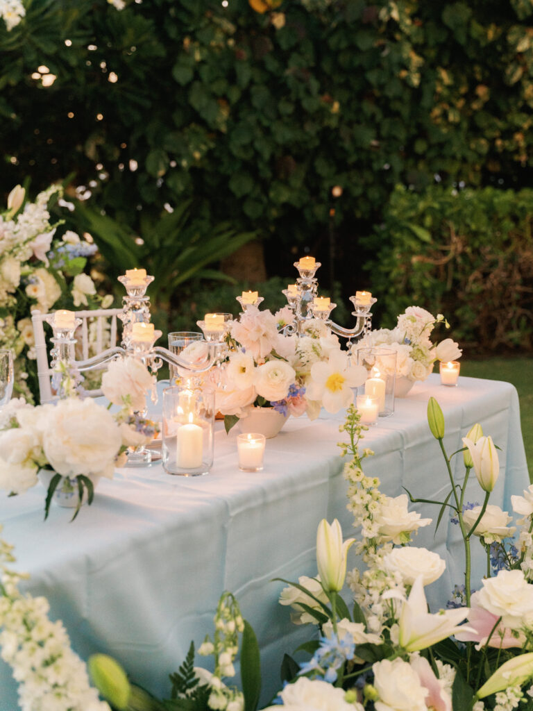 Luxury wedding reception table setting with light blue linens, lush white floral centerpieces, and glowing crystal candelabras outdoor