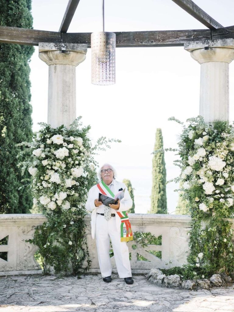 Elegant outdoor wedding ceremony featuring an officiant standing at a classic stone rotunda altar adorned with luxury white floral arrangements