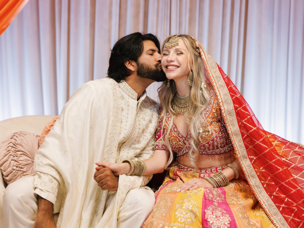 A bride and groom in traditional South Asian wedding attire share a candid, affectionate moment during their vibrant multicultural and traditional celebration.