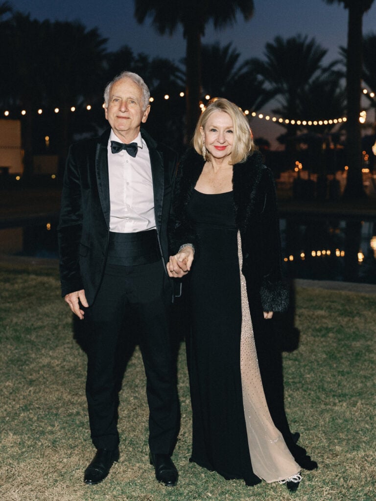 Wedding guests in black-tie attire including a classic tuxedo and sophisticated black gown at an outdoor evening venue