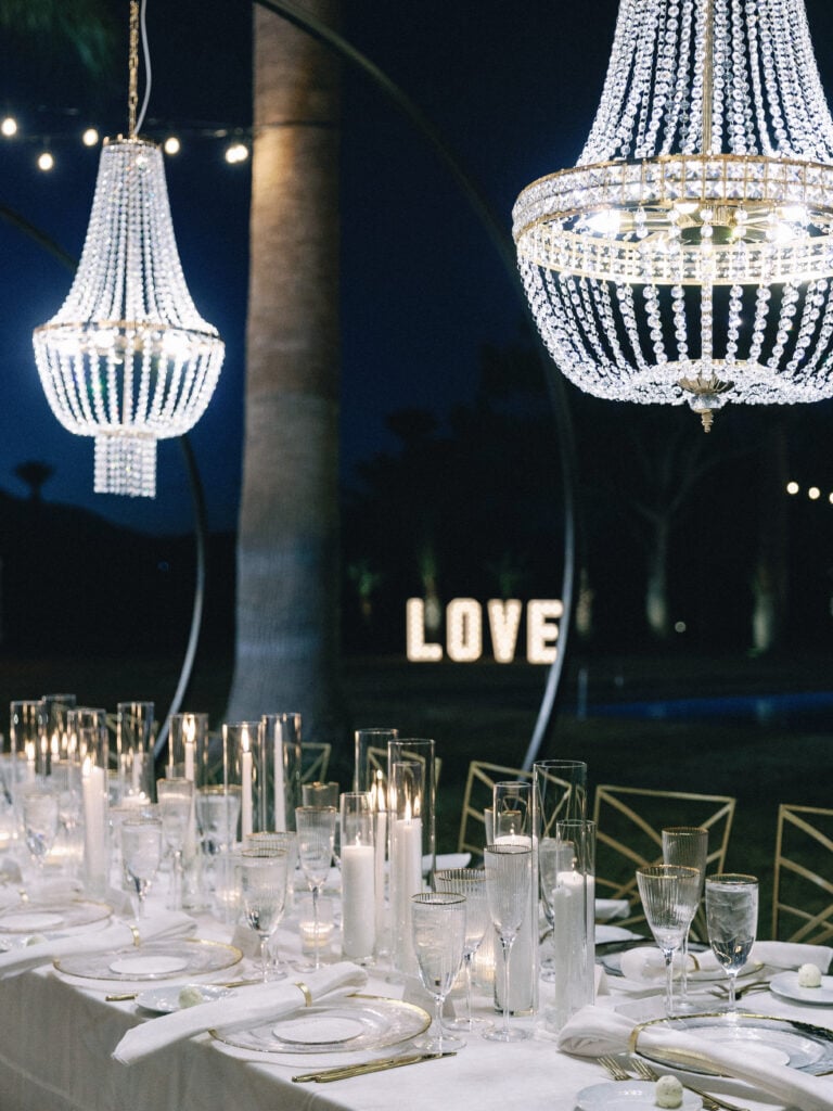 Luxury night wedding reception featuring long tables covered in white linens, candles, and hanging crystal chandeliers