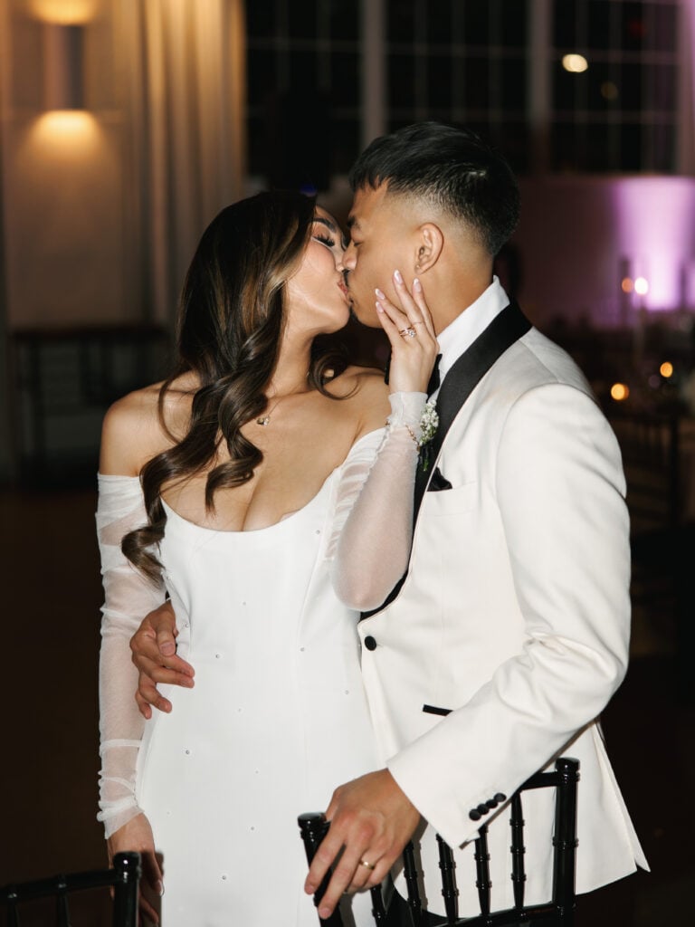 Bride in an off-the-shoulder wedding gown kissing the groom in a white tuxedo jacket