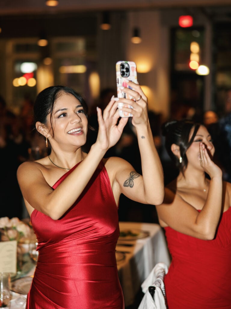 Smiling wedding guests in elegant red dresses taking photos and videos with a smartphone during the reception party