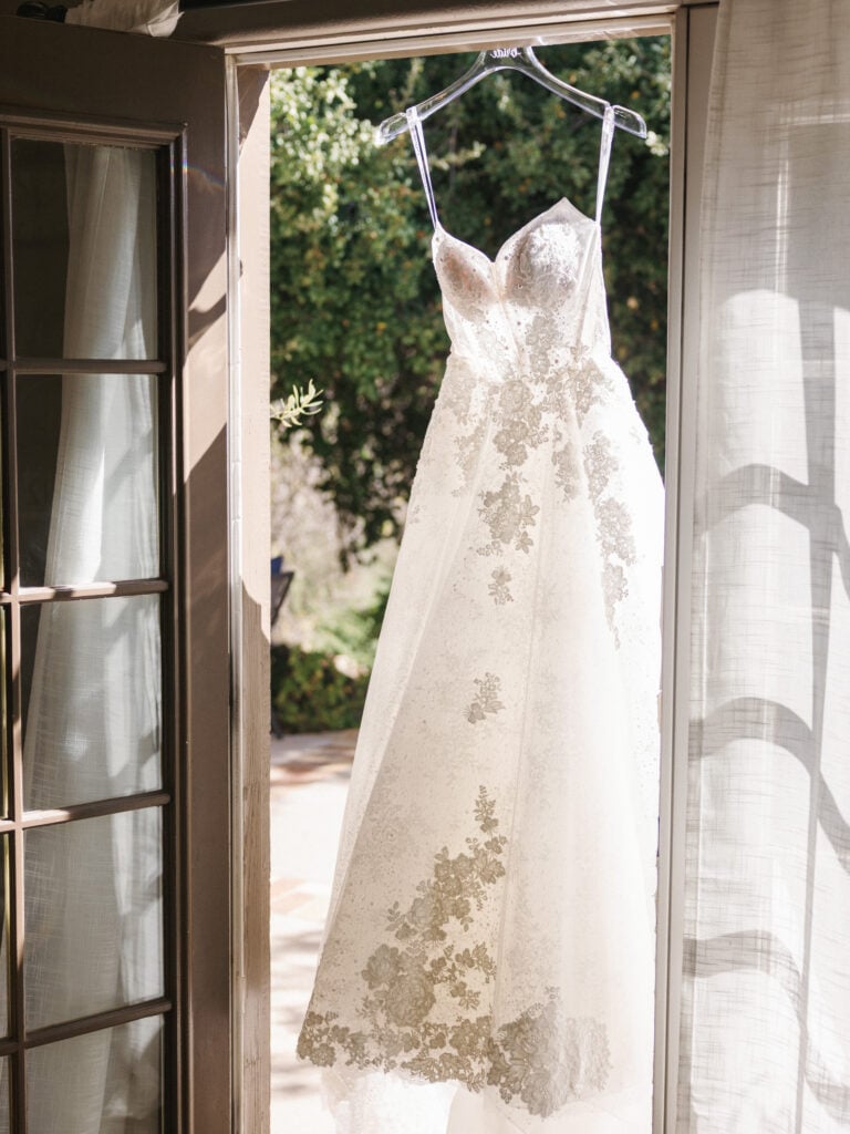 Strapless A-line wedding dress with intricate lace appliqués and an embroidered corset bodice hanging on a personalized acrylic hanger in a sunlit doorway during luxury morning bridal preparations