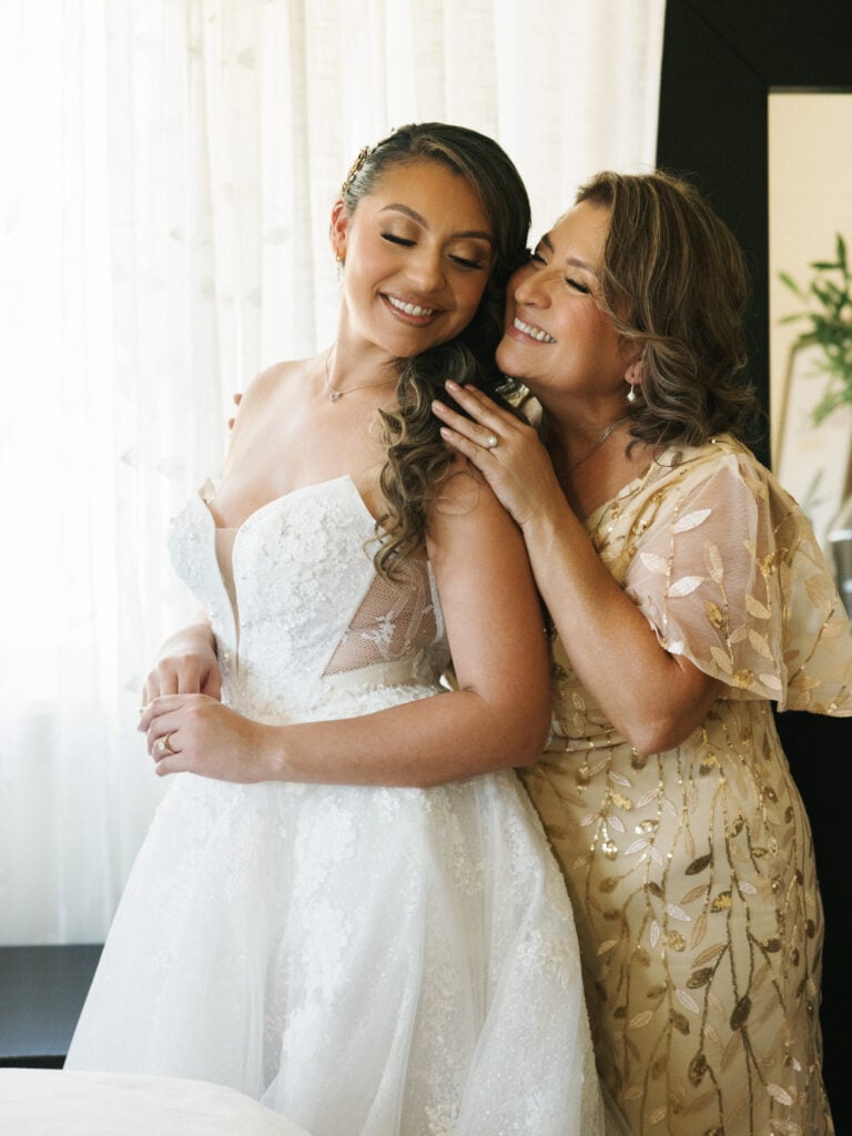Emotional mother of the bride embracing her daughter in a strapless embroidered lace wedding gown during morning preparation