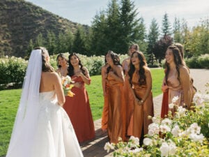 Joyful bridesmaids in terracotta satin gowns reacting with surprise during the bride’s first look reveal on a sunny garden lawn