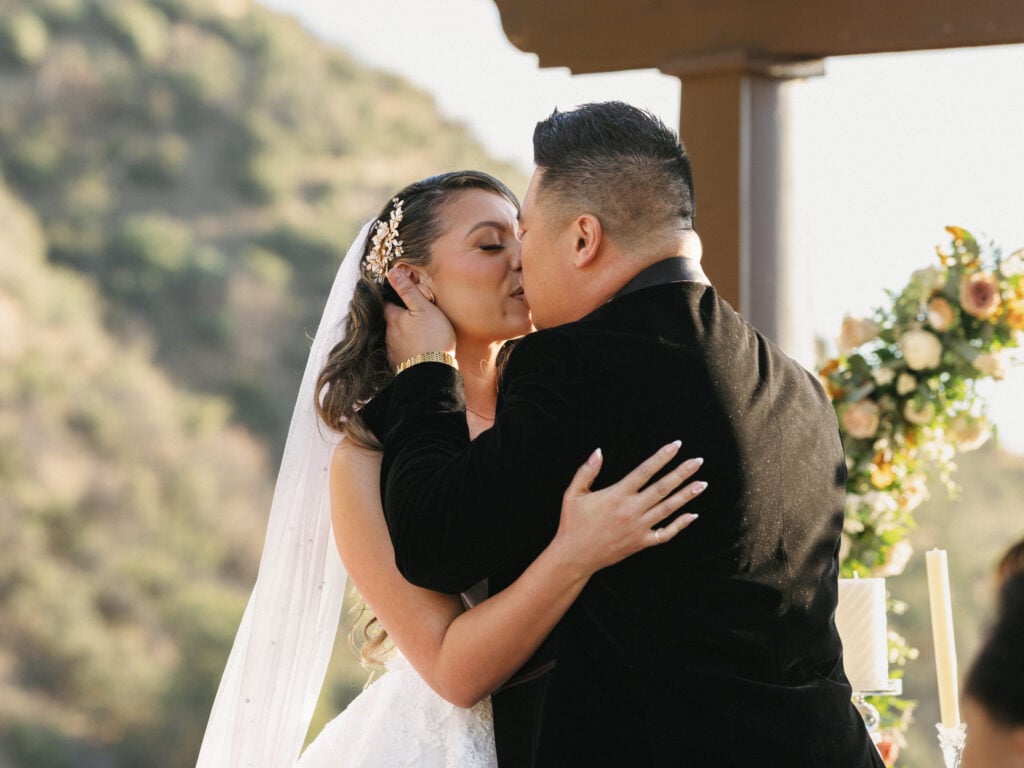 Groom in a black velvet tuxedo kissing his bride under a white veil at an outdoor luxury wedding ceremony with mountain view