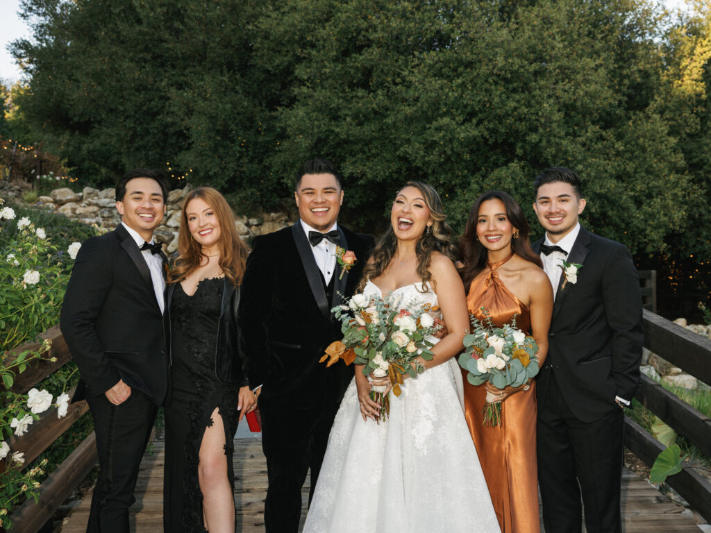 Bride in an embroidered gown and groom in a black velvet tuxedo celebrating with bridesmaids in terracotta and black dresses and groomsmen in classic tuxedos