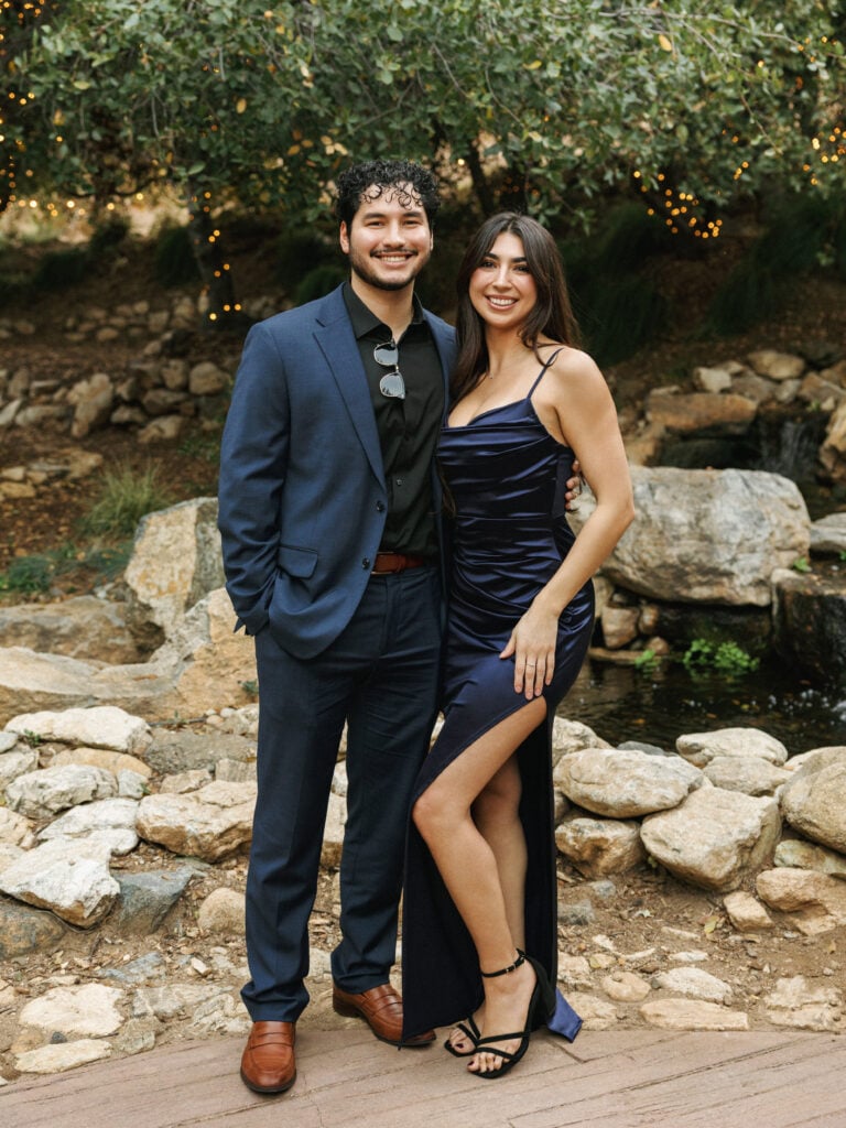 Elegant couple showcasing luxury wedding guest attire featuring a navy blue satin slit dress and a tailored blue suit at an upscale outdoor wedding reception