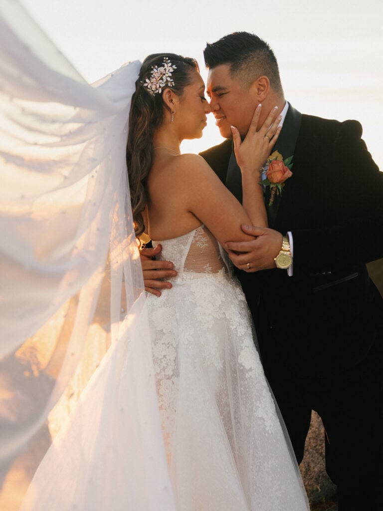 Groom in a black velvet tuxedo embracing his bride under a blowing white veil with pearls at sunset golden hour