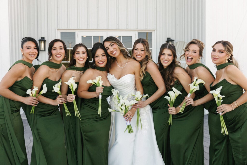 Bride in a strapless wedding dress with bridesmaids in emerald green satin gowns holding white calla lily bouquets