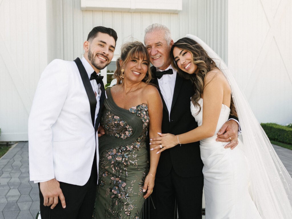 Bride in a veil and groom in a white tuxedo jacket smiling with family members in formal attire