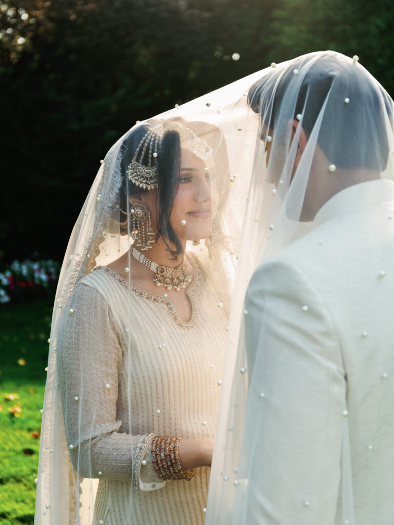 Traditional South Asian wedding couple photoshoot featuring a bride and groom under a pearl-embellished veil at sunset.
