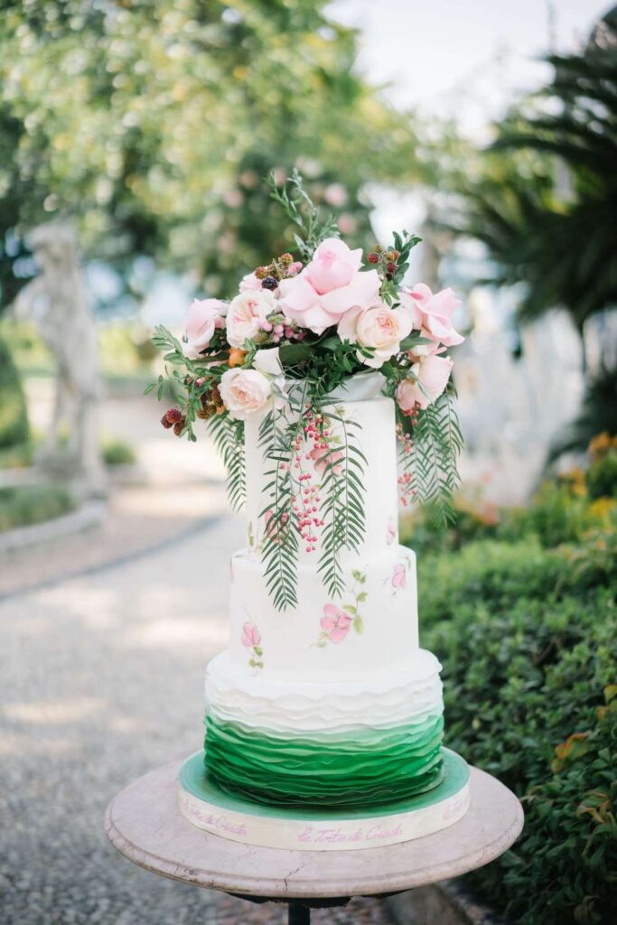 Elegant tiered wedding cake with green ruffle texture, hand-painted details, and a cascade of pink roses.