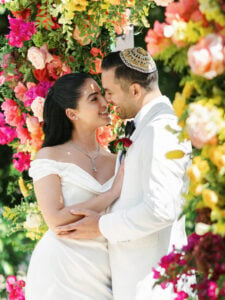 An upscale editorial capture of a luxury Jewish wedding, featuring the newlyweds embracing affectionately against a backdrop of breathtaking, vibrant floral chuppah designs.