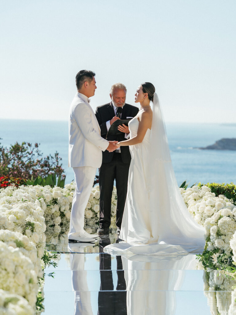 Professional wedding photography of a couple at the altar with a scenic ocean view and mirror aisle with white flowers.