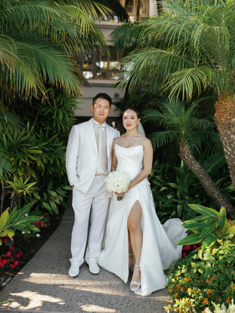 Full-length wedding portrait photography of a beautiful couple in white attire during an outdoor photoshoot in a lush garden.