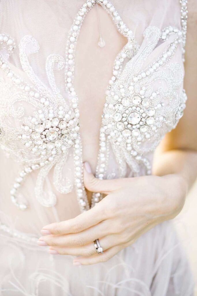 Couture wedding dress details featuring an intricate beaded bodice, pearl embellishments, and luxury diamond bridal rings close up.