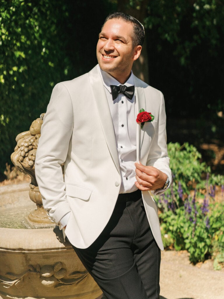 Groom in a white tuxedo jacket with a black bow tie and red rose boutonniere smiling outdoors