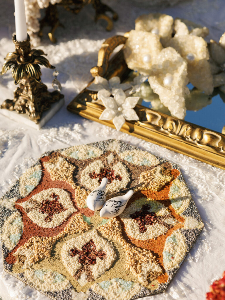A traditional Persian wedding display with intricate spice patterns and ceramic birds rests on a lace tablecloth during a Sofreh Aghd ceremony.