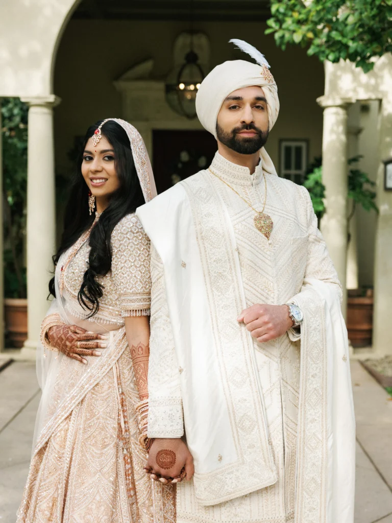A bride in a heavily beaded ivory lehenga stands back-to-back with her groom during their traditional Indian wedding celebration.