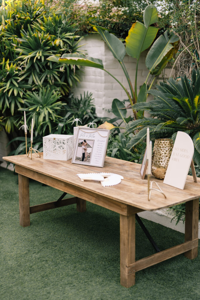 Outdoor wedding welcome table setup on a green lawn featuring a framed engagement photo, white guest book box, paper fans, and lush tropical greenery backdrop.