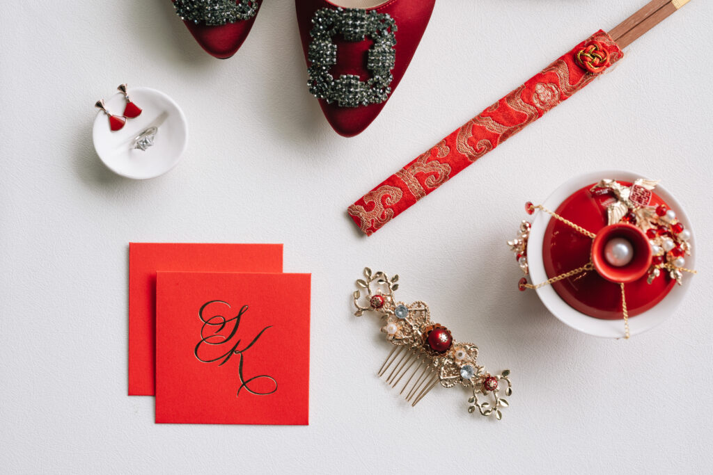 Red satin Manolo Blahnik Hangisi pumps with crystal buckles styled on a white surface with traditional Chinese wedding heirloom details, custom gold foil calligraphy envelopes, and a gold bridal hair comb for cultural wedding detail photos.