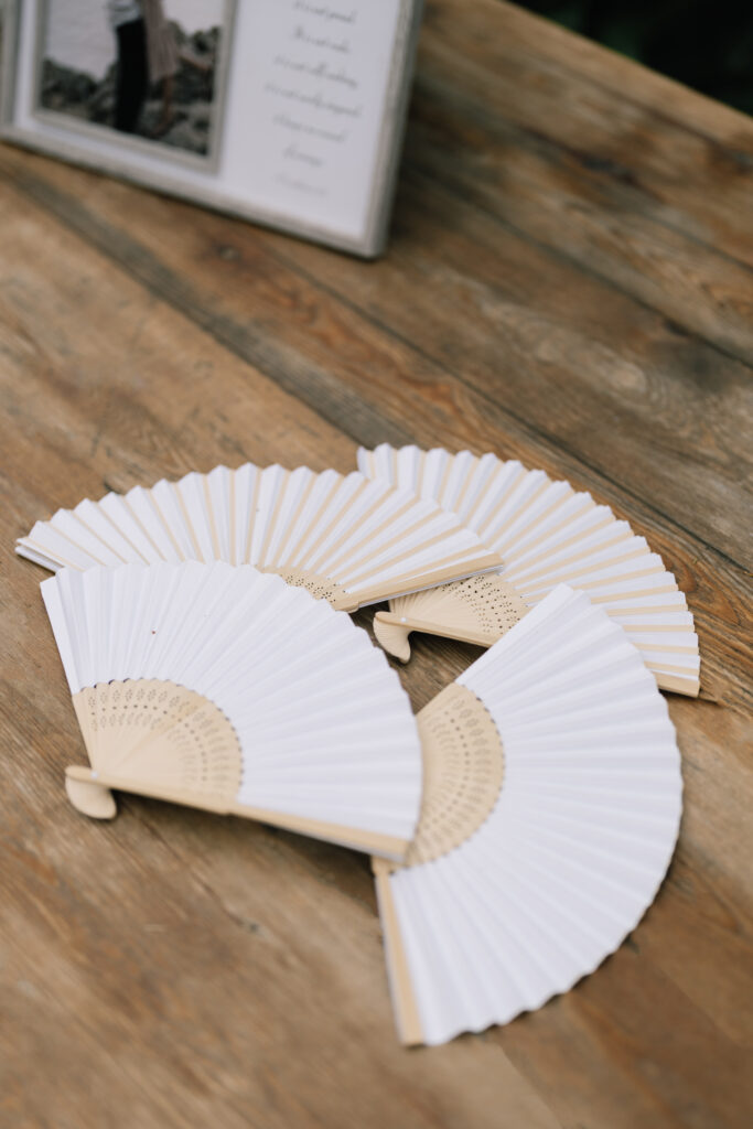 White paper folding fans with bamboo wood handles arranged on a rustic wooden welcome table for an outdoor summer wedding guest comfort.
