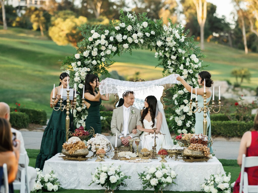 Editorial wedding photography of a Persian Sofreh Aghd ceremony showing a bride and groom under a floral arch during a multicultural wedding celebration.