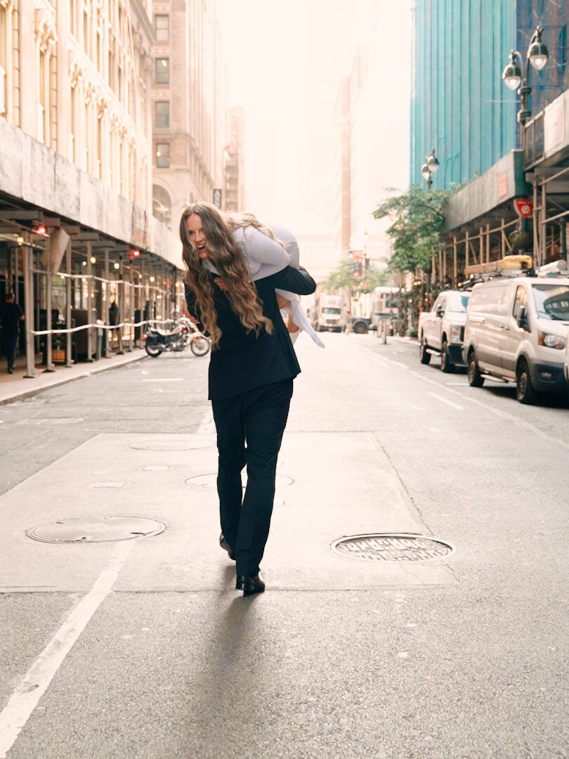 Candid city style wedding photography of a laughing woman in a dark suit giving a bride a fireman's carry down an urban street in New York.