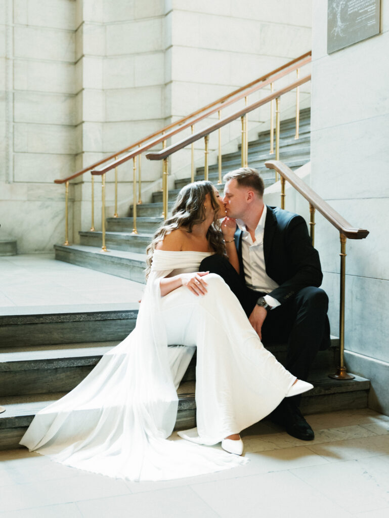 A bride and groom kiss while sitting on the historic marble staircase inside the New York Public Library wedding venue.