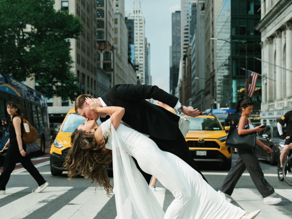 Newlyweds kissing in a crosswalk as city traffic moves behind them.
