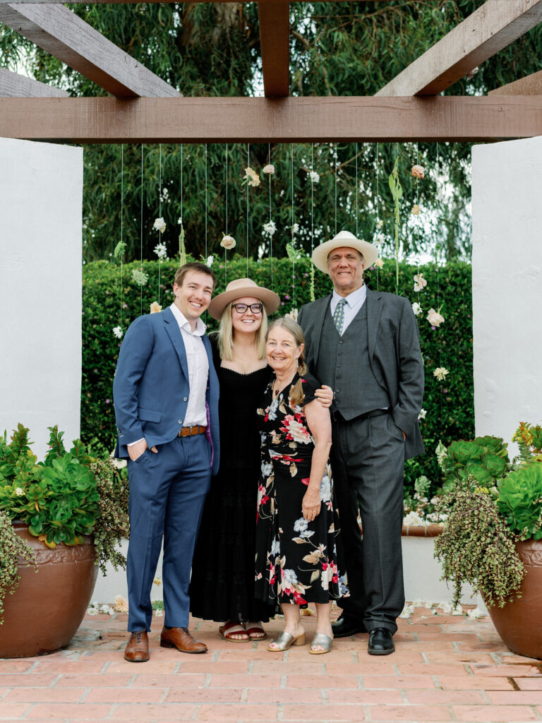 Sophisticated wedding guest and parents photo in front of a modern hanging floral installation on a terracotta tile patio at a luxury California estate.