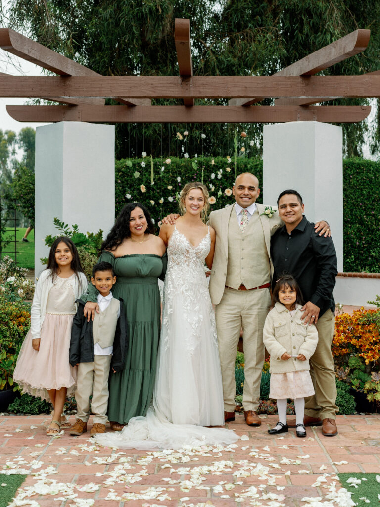 Candid family wedding portrait under a wooden pergola, showcasing a modern bride and groom in a beige suit smiling with their relatives and children on a flower-petal covered patio.