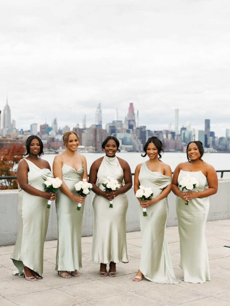 Bridesmaids in elegant sage green satin dresses hold white rose bouquets with the Manhattan skyline in the background.