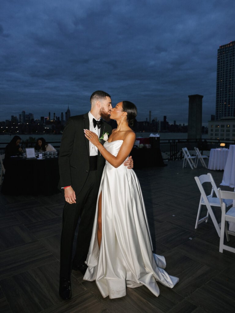 Bride in a strapless satin wedding dress with a high slit kissing a groom in a black tuxedo and bow tie on an outdoor rooftop terrace at dusk with the New York City skyline in the background.