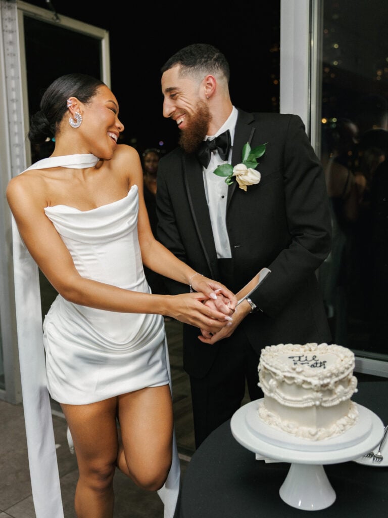 Medium shot of a wedding couple cutting a lambeth-style heart cake with cursive writing on top, showcasing the bride's modern mini wedding gown with a long neck scarf.