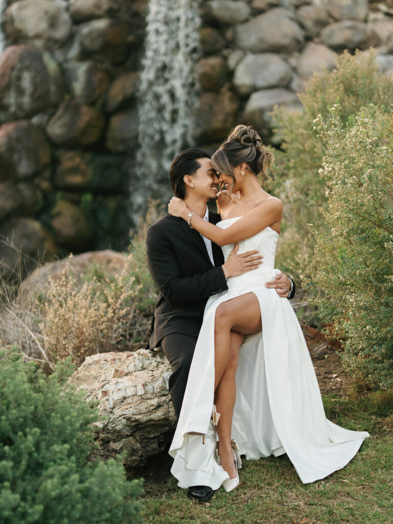 Romantic newlyweds posing on rocks by a garden waterfall, showcasing a contemporary strapless high-slit wedding gown and white bridal heels.