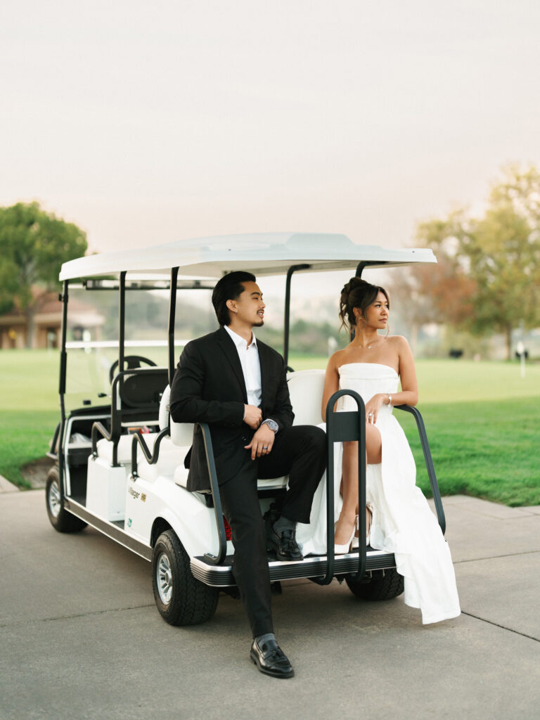 Bride in a strapless dress with a high slit sitting in a white golf cart while the groom in a black suit poses on a paved country club path.