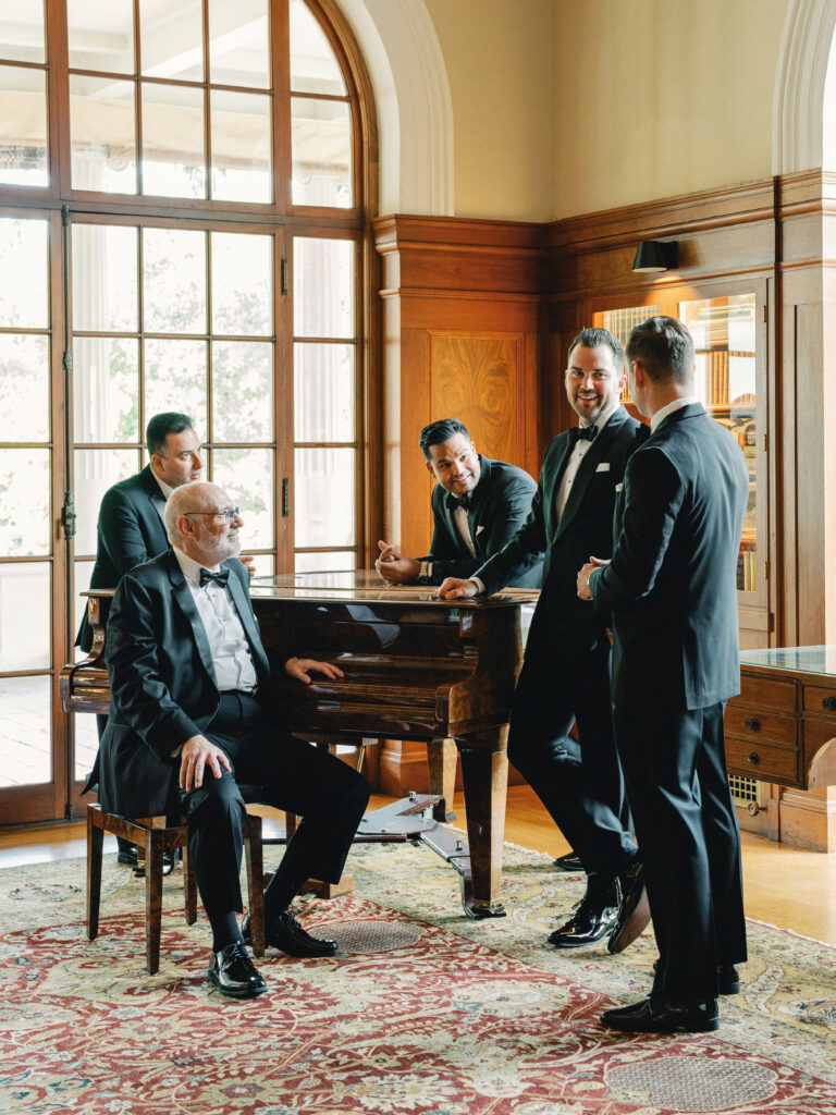 Groom and groomsmen in black tuxedos talking with a senior family member seated at a grand piano inside a luxury wood-paneled estate room.