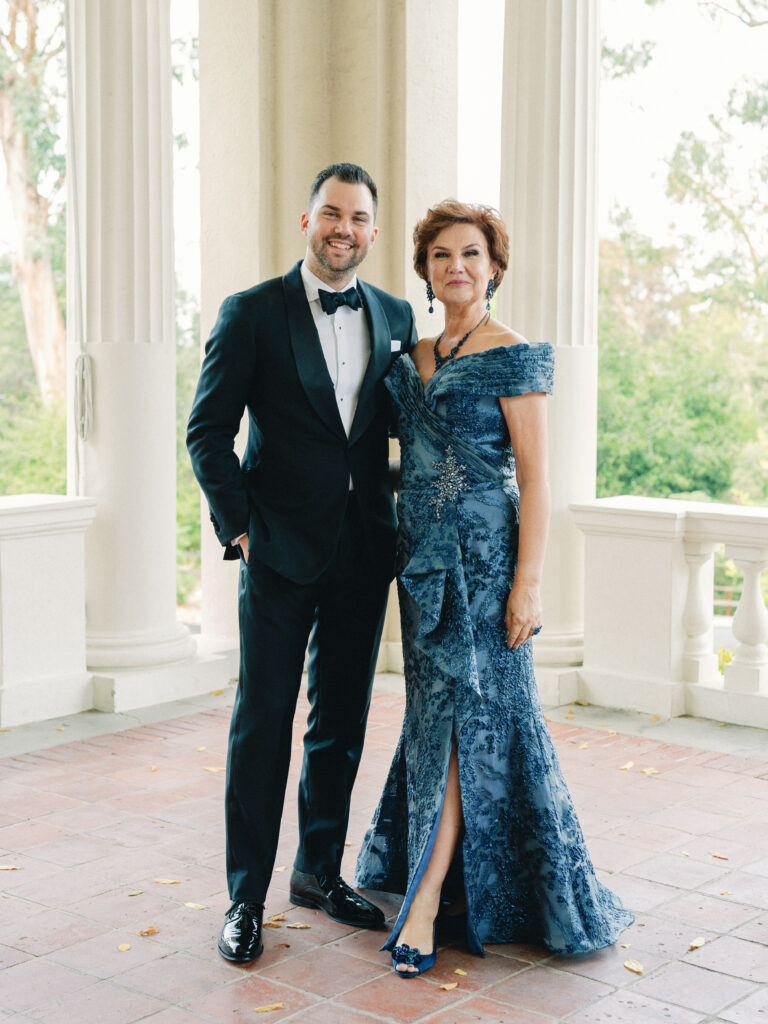 Sophisticated Mother of the Groom portrait on a luxury estate terrace with white pillars, featuring a handsome groom in a black tuxedo and his mother in an elegant navy blue lace gown.