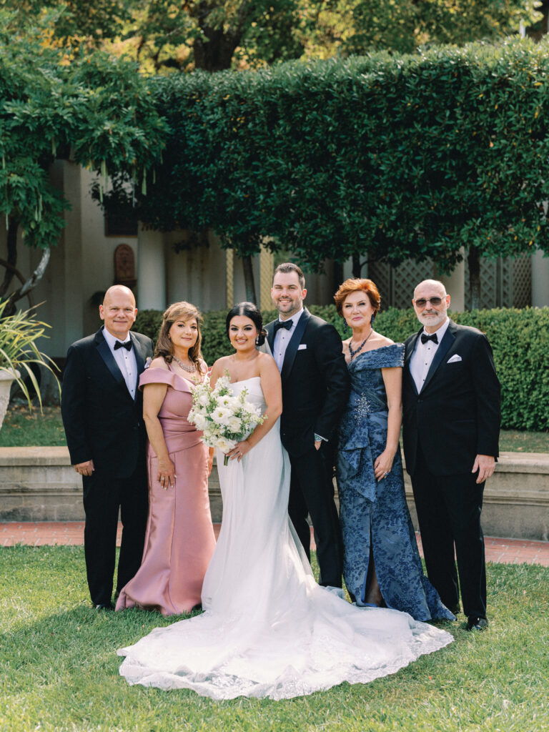 Elegant wedding family portrait with the bride, groom, and their parents in formal attire outdoors.