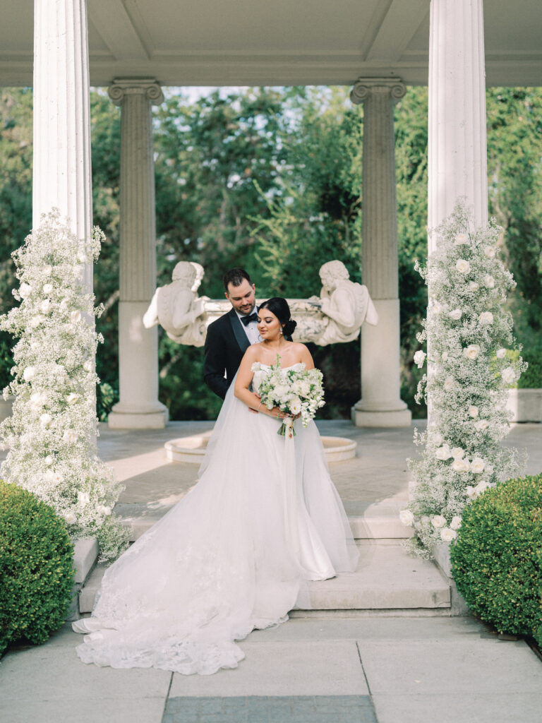 A bride and groom pose by a historic stone rotunda with white rose and baby's breath floral installations at a luxury Santa Barbara estate wedding.
